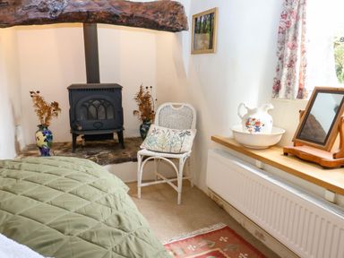 A bedroom corner with a wood burning stove, a white wicker chair with a cushion, a decorative jug and bowl on a wooden shelf, and floral curtains at Bridge Cottage in Dalwood