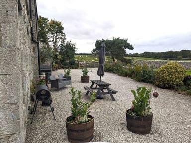 An outdoor patio with gravel ground, wooden picnic table, closed umbrella, two planters with plants, and a stone wall in the background at The Dairy at Brackenthwaite Farm in Arnside