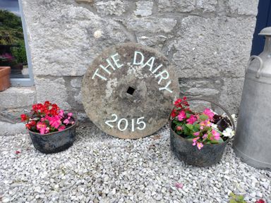 A stone wall with a round stone sign reading The Dairy 2015 between two metal baskets with flowers and a metal milk canister on gravel at The Dairy at Brackenthwaite Farm in Arnside
