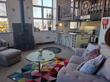 A living room with a fireplace a colorful rug a glass coffee table and a kitchen area with bar stools at The Dairy at Brackenthwaite Farm Arnside
