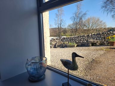 A window view showing a gravel yard with stone wall and flower pots and a wooden bird decoration and a blue glass bowl on the window sill at The Dairy at Brackenthwaite Farm in Arnside