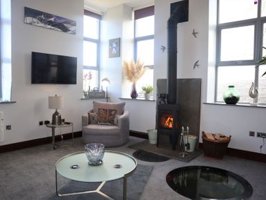 A living room with a wood stove an armchair a round coffee table and a wall-mounted TV at The Dairy at Brackenthwaite Farm Arnside
