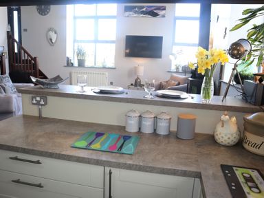 A kitchen countertop with containers labeled tea sugar coffee a bread box and a vase of yellow flowers with a living area and windows in the background at The Dairy at Brackenthwaite Farm in Arnside