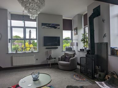 A living room with a round chair a wall-mounted TV a wood-burning stove and windows at The Dairy at Brackenthwaite Farm Arnside