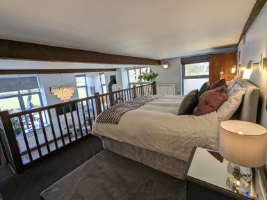 A bedroom with a large bed and bedside table overlooking a living room with windows at The Dairy at Brackenthwaite Farm in Arnside