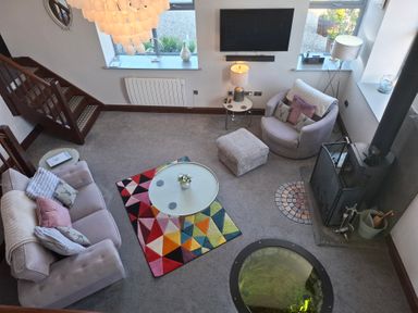 A living room with a sofa, armchair, round glass coffee table, colorful rug, wood stove, and two windows at The Dairy at Brackenthwaite Farm in Arnside