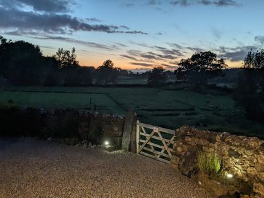 A gravel driveway with a wooden gate and stone wall overlooking a field with trees at sunset at The Dairy at Brackenthwaite Farm in Arnside