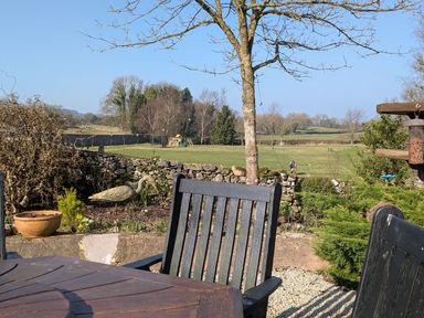 An outdoor seating area with wooden table and chairs overlooking a garden with a stone wall and a grassy field at The Dairy at Brackenthwaite Farm in Arnside