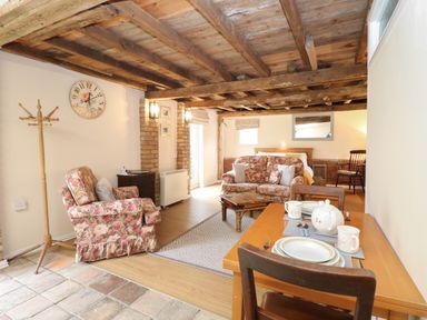 A living area with floral armchair and sofa a wooden dining table set with dishes a bed in the background and a wooden beamed ceiling at Pear Tree Barn in Stalham