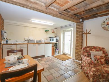 A kitchen area with cabinets a dining table set with dishes a floral armchair a coat rack and a door at Pear Tree Barn in Stalham