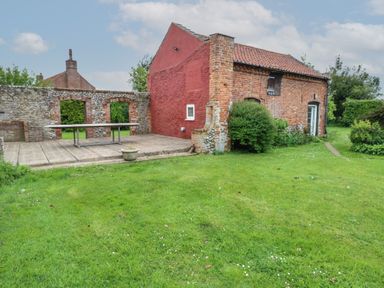 A brick building with a red wall and tiled roof next to a stone wall and patio with a table on a lawn at Pear Tree Barn in Stalham
