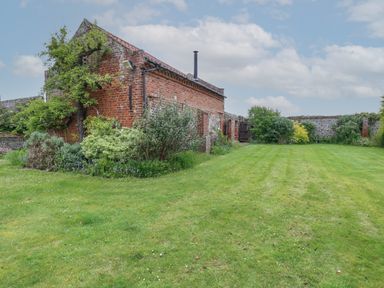 A grassy garden next to a brick building with plants and a wooden gate at Pear Tree Barn in Stalham