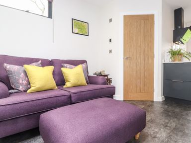 A living room with a purple sofa and ottoman yellow and floral cushions a wooden door and a black kitchen unit with a plant at Yew Tree Cabin in Ullswater