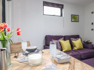 A dining table set with glasses and bowls next to a purple sofa with yellow and patterned cushions in a living area at Yew Tree Cabin in Ullswater