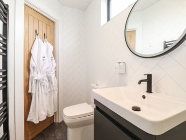 A bathroom with a white sink and black faucet a round mirror a toilet and white robes hanging on a wooden door at Yew Tree Cabin in Ullswater