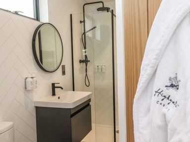 A bathroom with a black sink faucet round mirror walk-in shower with black frame and white robe hanging on door at Yew Tree Cabin in Ullswater