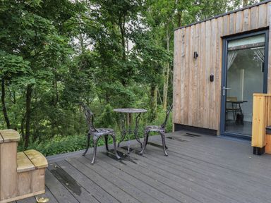 A wooden deck with two metal chairs and a small round table outside a building surrounded by trees at Yew Tree Cabin in Ullswater