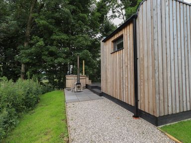 A wooden cabin exterior with a gravel path and patio area with a hot tub stove beside trees at Yew Tree Cabin in Ullswater