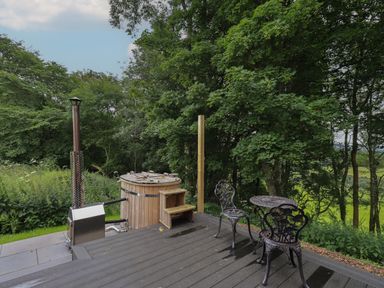 An outdoor wooden hot tub and a metal table with two chairs on a wooden deck surrounded by trees at Yew Tree Cabin in Ullswater