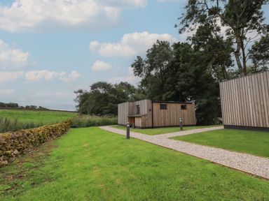 A grassy area with a gravel pathway between wooden buildings and trees in the background at Yew Tree Cabin in Ullswater