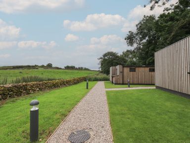 A gravel path with modern lamp posts running through a grassy area next to wooden buildings and a stone wall at Yew Tree Cabin in Ullswater
