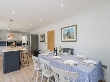 A dining area with a blue patterned tablecloth on a table set with white plates and glasses next to a kitchen with an island and three stools at Greystones in Upton St Leonards
