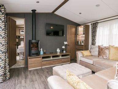 A living room with a wall-mounted tv a wood stove beige sectional sofa and a cabinet with glassware at 43 Pony Meadow in Troutbeck Bridge
