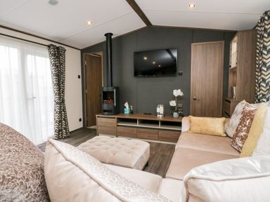 A living room with a beige sectional sofa a tufted ottoman a wall-mounted television a wood-burning stove and patterned curtains at 43 Pony Meadow in Troutbeck Bridge