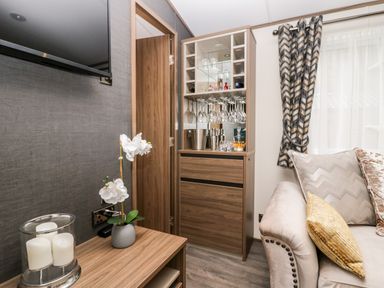 A living room corner with a wooden cabinet holding glasses and bottles a sofa with pillows a small table with a flower pot and candles and a mounted TV at 43 Pony Meadow in Troutbeck Bridge