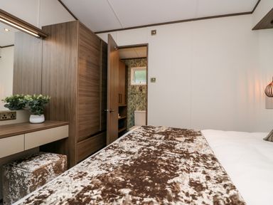 A bedroom with a bed a brown and white blanket a wooden wardrobe a dressing table and a door leading to a bathroom at 43 Pony Meadow in Troutbeck Bridge