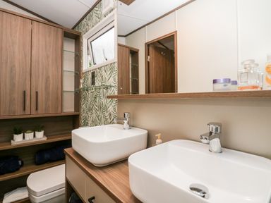 A bathroom with two white sinks on a wooden countertop a wooden cabinet with shelves a toilet and a window with tropical leaf wallpaper at 43 Pony Meadow in Troutbeck Bridge