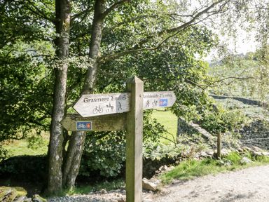 A wooden signpost with directions to Grasmere and Ambleside on a gravel path surrounded by trees and stone walls at 43 Pony Meadow in Troutbeck Bridge