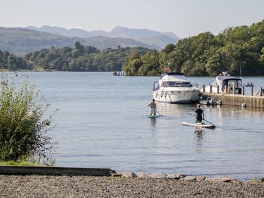 Two people paddleboarding on a lake near boats docked at a pier with trees and mountains in the background at 43 Pony Meadow in Troutbeck Bridge