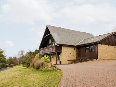 Exterior view of a house with a stone and wood facade and a sloped roof with a driveway and grass lawn at Slaley Park Villa in Hexham