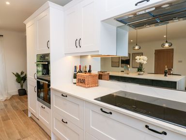 A kitchen with white cabinets an induction cooktop built in oven bottles and a basket on the countertop and a plant in the corner at Slaley Park Villa in Hexham