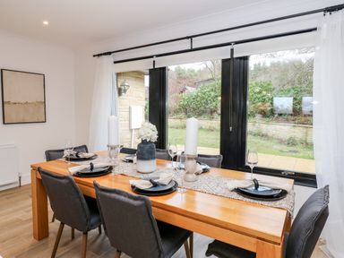 A dining room with a wooden table set with plates candles and flowers and black chairs by a large window with white curtains at Slaley Park Villa in Hexham