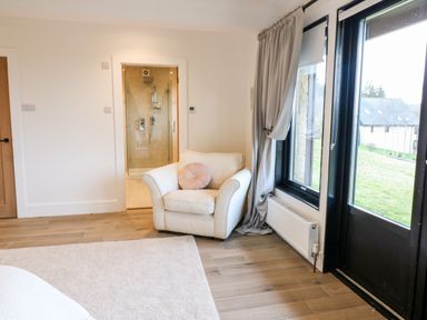 An armchair with a round pillow next to a glass shower door and large windows with curtains at Slaley Park Villa in Hexham