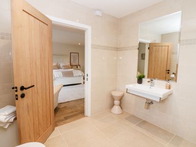 A bathroom with beige tiled walls and floor a white sink with a mirror above a beige stool and an open wooden door showing a bedroom at Slaley Park Villa in Hexham