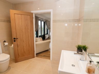 A bathroom with a white sink a plant a soap dispenser a toilet and an open wooden door showing a living room with a beige sofa and windows with curtains at Slaley Park Villa in Hexham