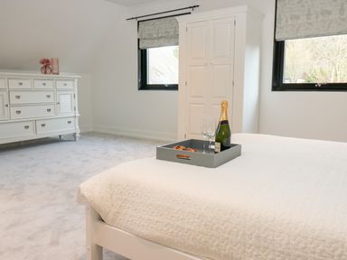A bedroom with a white bed covered by a quilt and a tray holding a bottle of champagne and two glasses a white dresser with flowers on it and a white wardrobe are near two windows with patterned blinds at Slaley Park Villa in Hexham