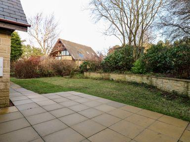 A backyard with a tiled patio and grass lawn surrounded by bushes and trees with a house in the background at Slaley Park Villa Hexham