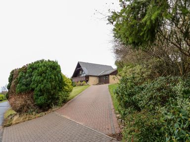 A house with a sloped roof at the end of a paved driveway surrounded by bushes and trees at Slaley Park Villa in Hexham