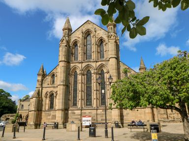 A stone church with arched windows and pointed towers beside a tree and benches with people sitting at Slaley Park Villa in Hexham