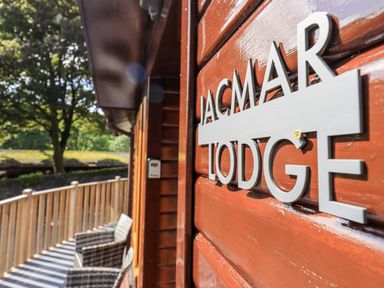 A wooden lodge exterior with a sign reading JacMar Lodge and outdoor chairs on a balcony surrounded by trees at JacMar Lodge 14 Applethwaite Limefitt Park near Windermere