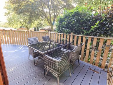 An outdoor wooden deck with a glass top table and four wicker chairs surrounded by a wooden fence and greenery at JacMar Lodge 14 Applethwaite Limefitt Park near Windermere