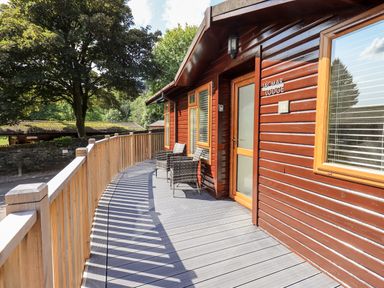 An outdoor wooden deck with two chairs and railing outside a lodge with trees in the background at JacMar Lodge 14 Applethwaite Limefitt Park near Windermere