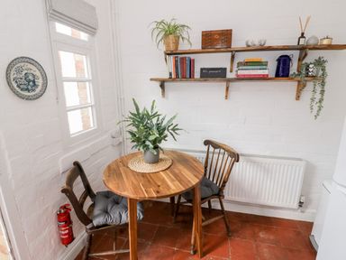 A small dining area with a round wooden table and two wooden chairs with cushions a potted plant on the table a white brick wall with wooden shelves holding books plants and decorative items a fire extinguisher near the window at Ross Cottage in Broadway