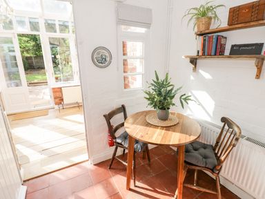 A small dining area with a round wooden table two chairs with cushions a plant on the table and a shelf with books and a wifi password sign at Ross Cottage in Broadway