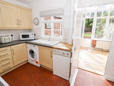 A kitchen with wooden cabinets a microwave toaster washing machine and a dishwasher with a door opening to a sunlit conservatory and garden at Ross Cottage in Broadway