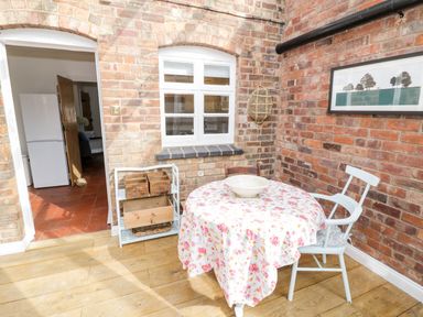 A dining area with a round table covered in a floral tablecloth two chairs wooden floor and exposed brick walls at Ross Cottage in Broadway